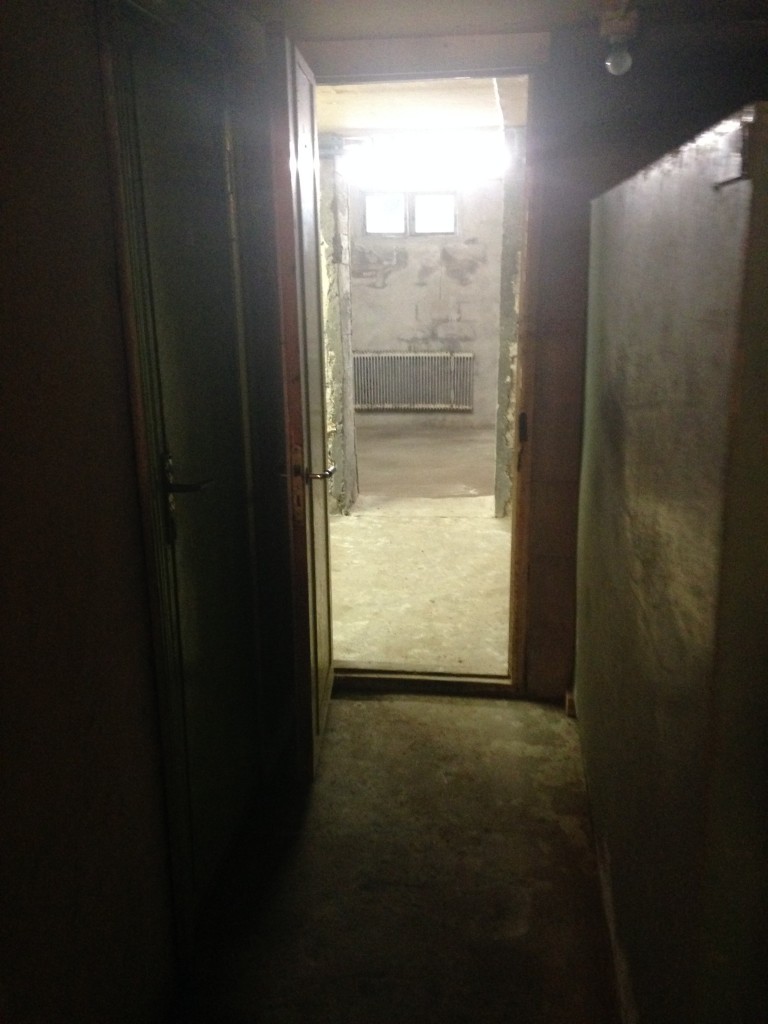 En korridor i källaren. Dörren till vänster leder in till toaletten, till höger står en gammal oljetank och rakt fram kommer garaget.